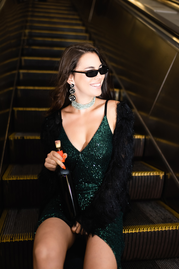 mulher glamour em óculos de sol sentado na escada rolante metro com garrafa de vinho - Foto, Imagem