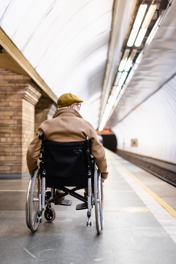 vista posteriore dell'anziano sulla sedia a rotelle, con cappotto e cappuccio, sulla piattaforma della stazione della metropolitana - Foto, immagini