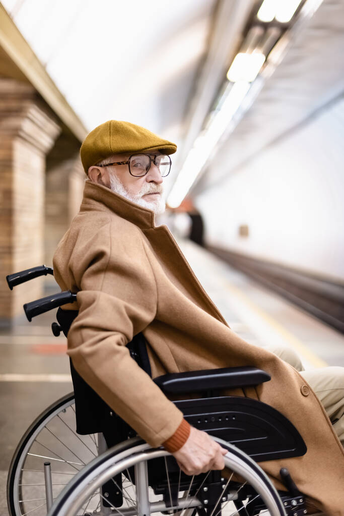 Sonbahar ceketli ve şapkalı yaşlı adam metro istasyonunda tekerlekli sandalyede oturuyor. - Fotoğraf, Görsel