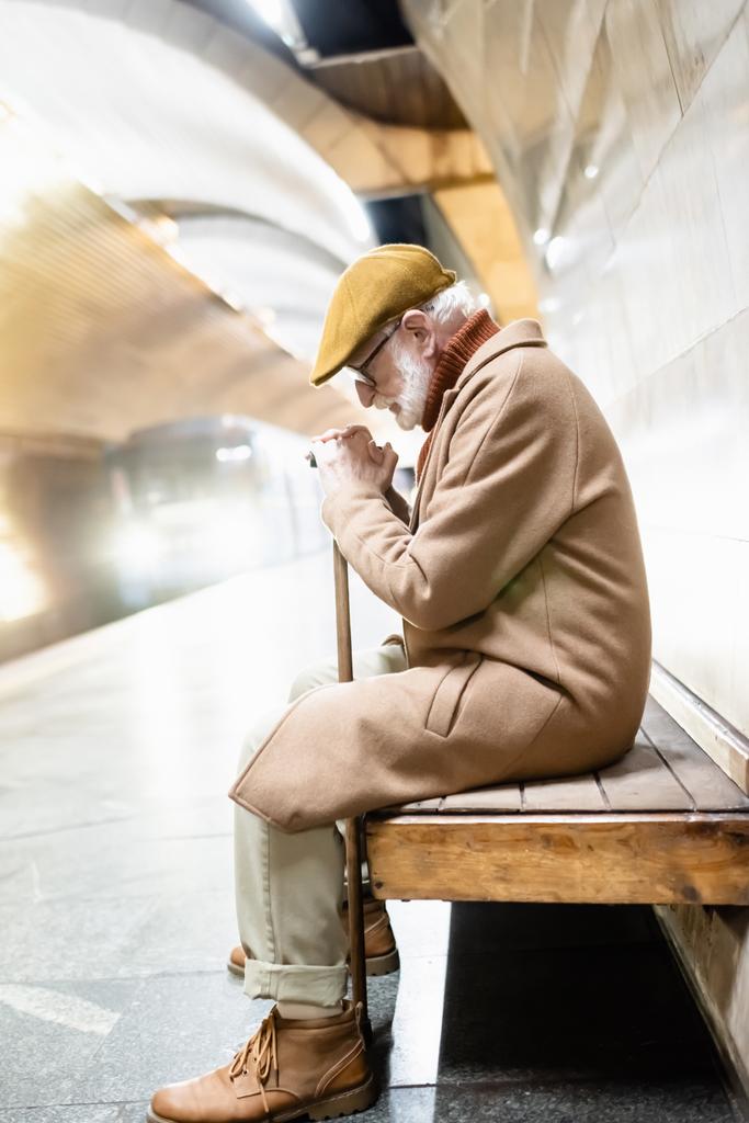 zijaanzicht van senior man zittend op bank met gebogen hoofd terwijl de trein aankomt op metroperron - Foto, afbeelding