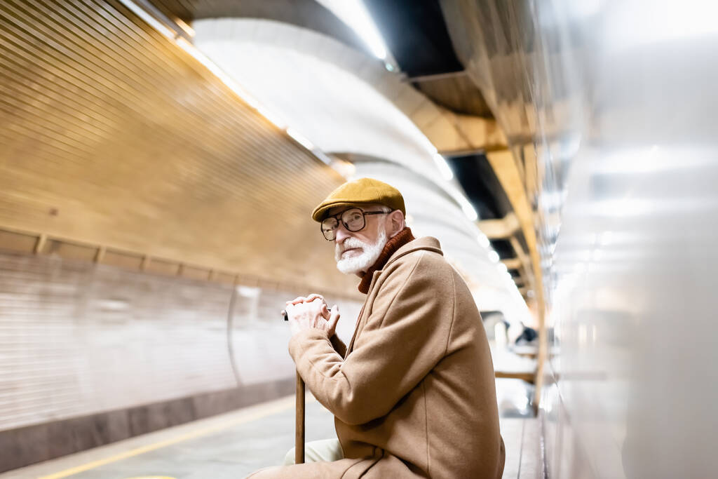 Sonbahar kıyafetli ve gözlüklü yaşlı adam elinde bastonla yeraltı platformunda oturuyor. - Fotoğraf, Görsel