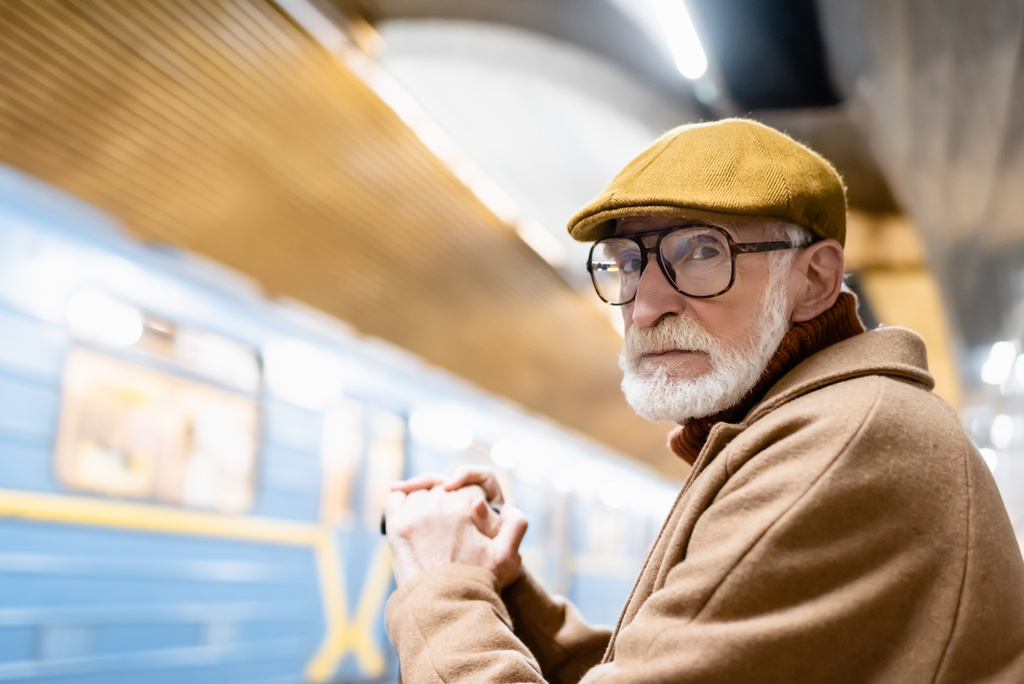 senior man in herfstkleding en bril kijkend naar camera op metroperron met wazige trein - Foto, afbeelding