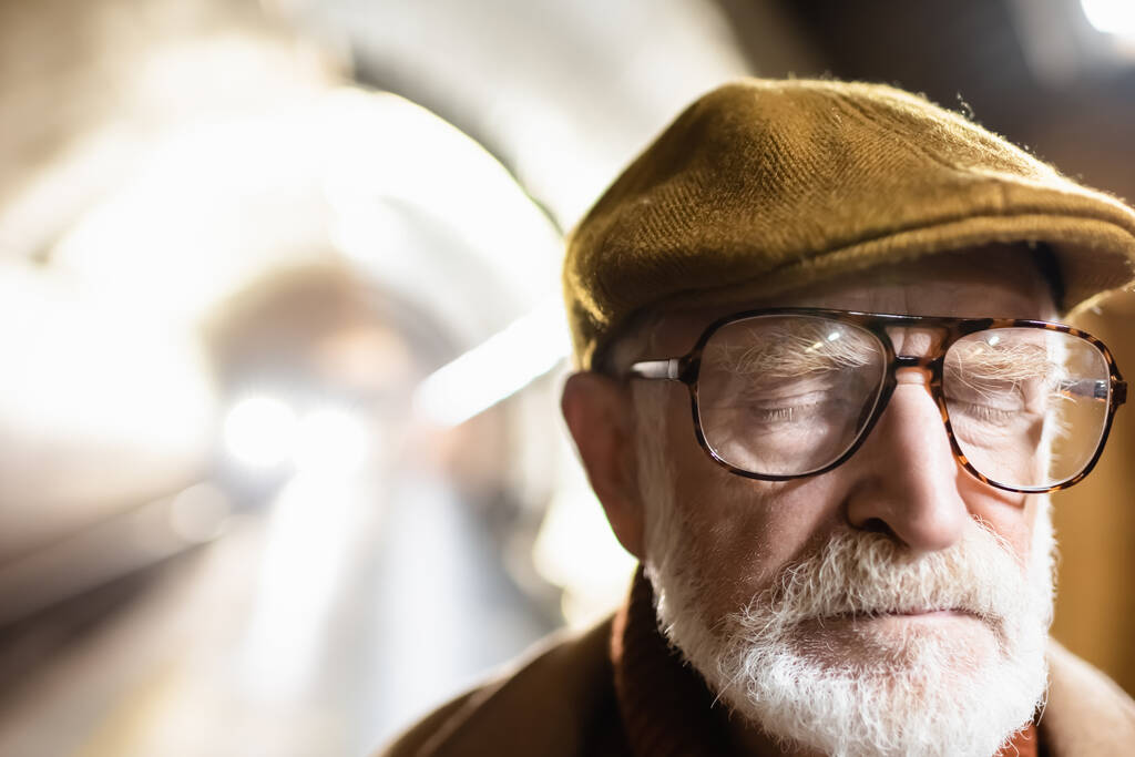 portrait of aged man in cap and eyeglasses standing with closed eyes on metro platform with light in metro tunnel on background - Fotografie, Obrázek