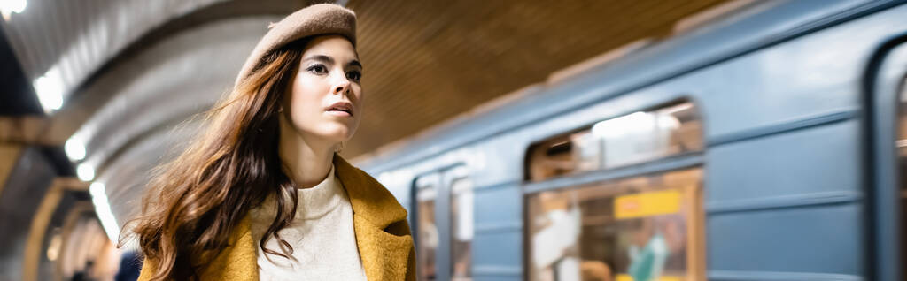 Güz beresi ve ceketli genç bir kadın metro istasyonunda bulanık trenin yanından uzağa bakıyor. - Fotoğraf, Görsel