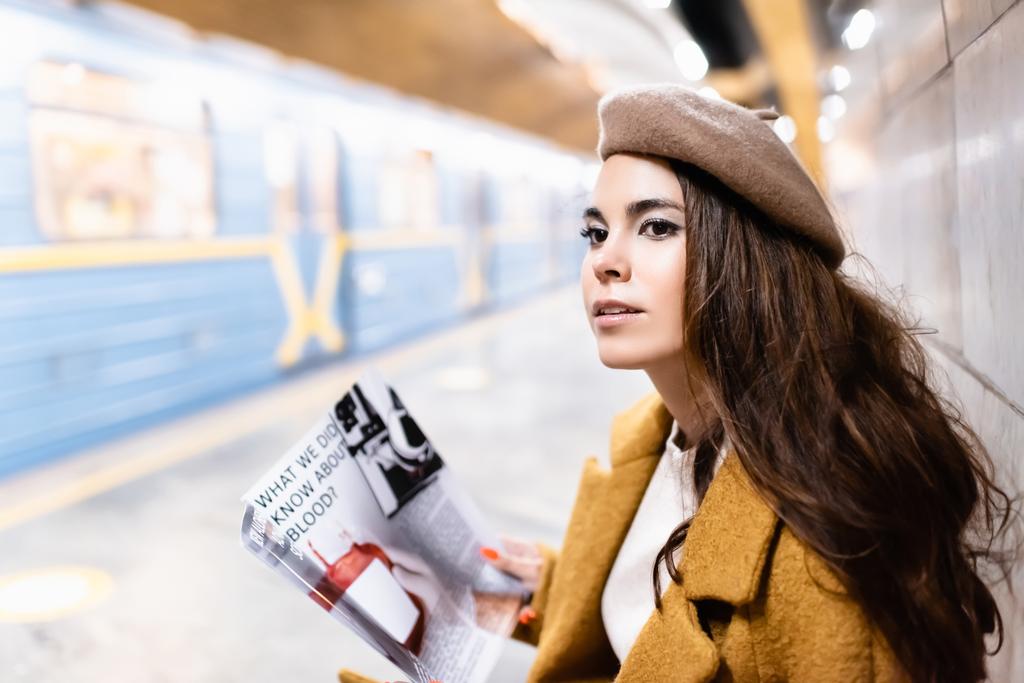 young woman in beret holding magazine while sitting on metro platform with blurred train - Photo, Image