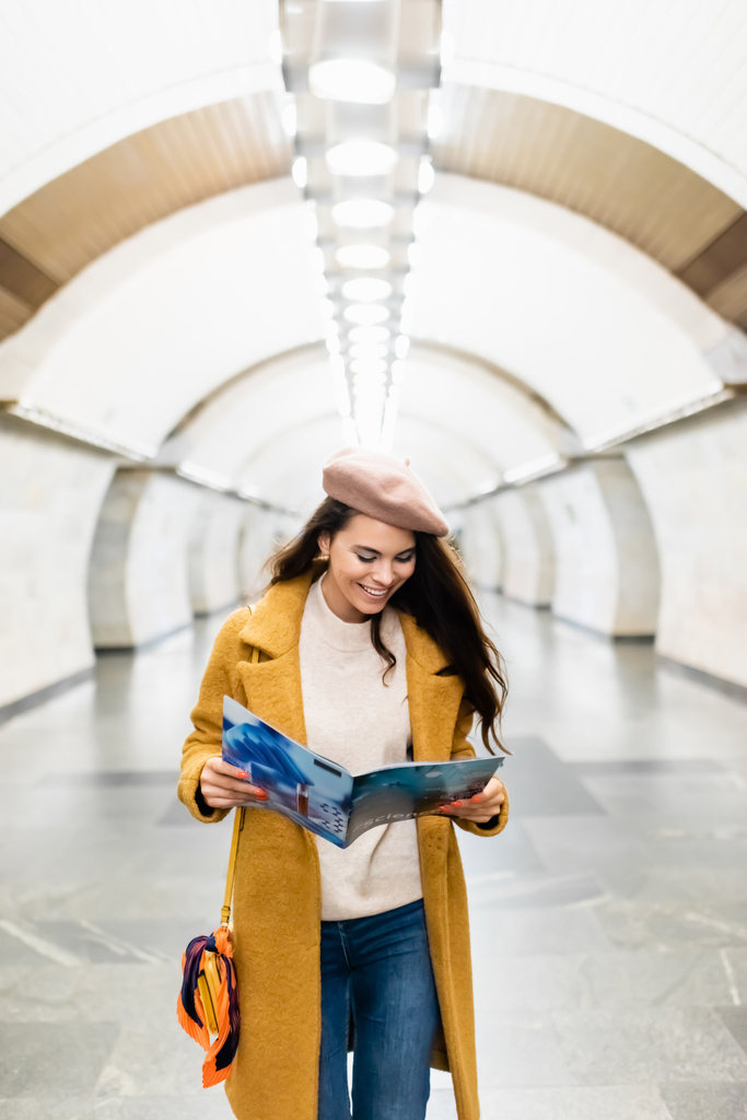cheerful woman in stylish autumn clothes reading magazine at subway station - Foto, Imagen