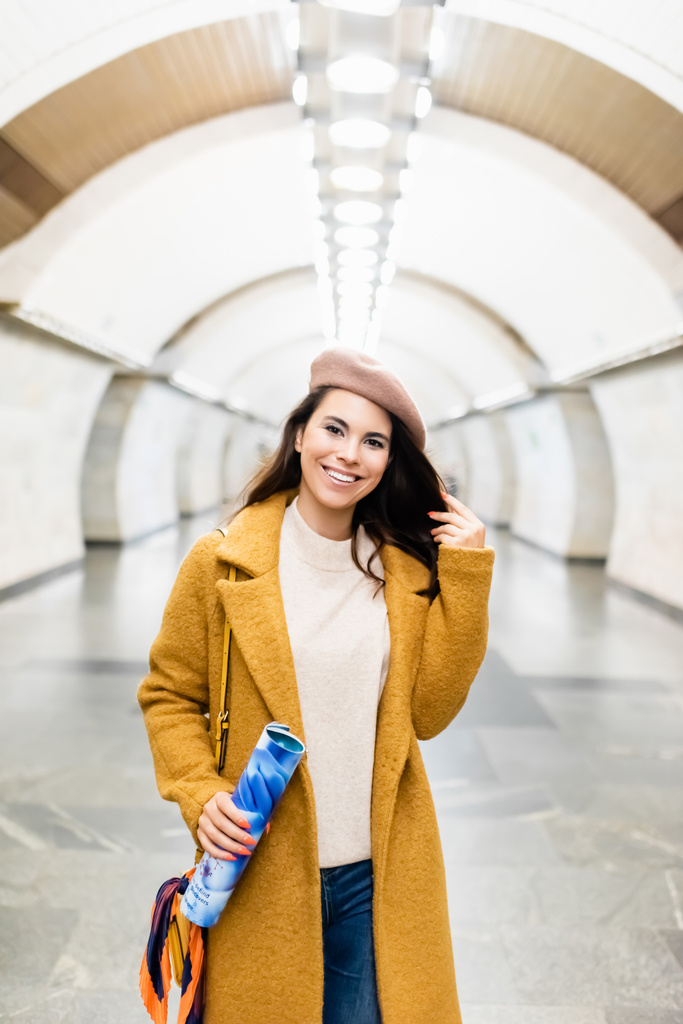 onnellinen, tyylikäs nainen takki ja baskeri katsot kameraa samalla kun pidät lehteä metroasemalla - Valokuva, kuva