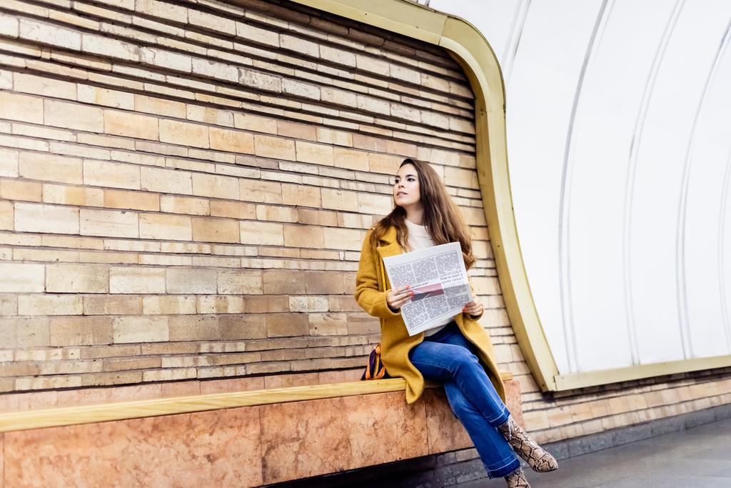 κομψή γυναίκα το φθινόπωρο ρούχα κοιτάζοντας μακριά, ενώ κάθεται στο παγκάκι πλατφόρμα του μετρό με εφημερίδα - Φωτογραφία, εικόνα