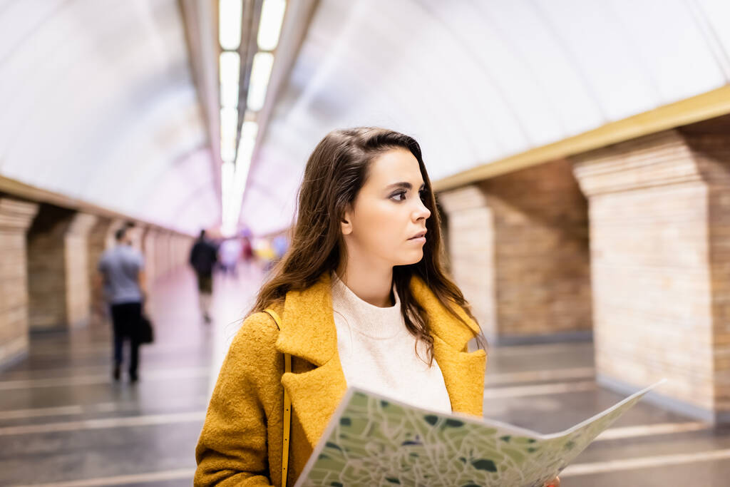 young woman in autumn coat looking away while holding city map at subway station - Fotografie, Obrázek