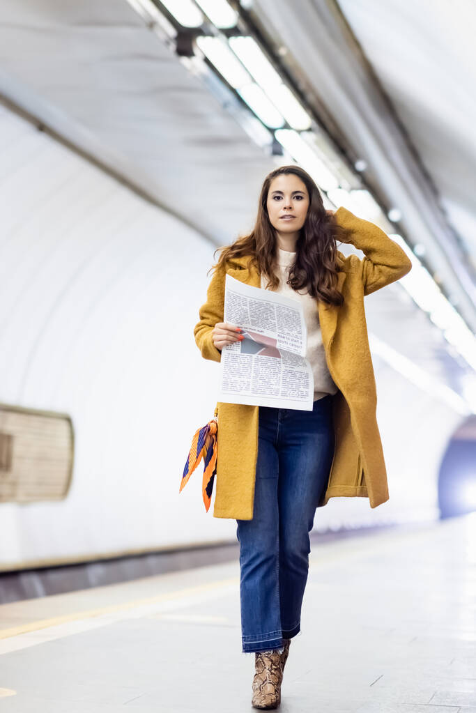 jonge vrouw in stijlvolle herfst outfit kijken naar camera terwijl het houden van krant op metrostation - Foto, afbeelding