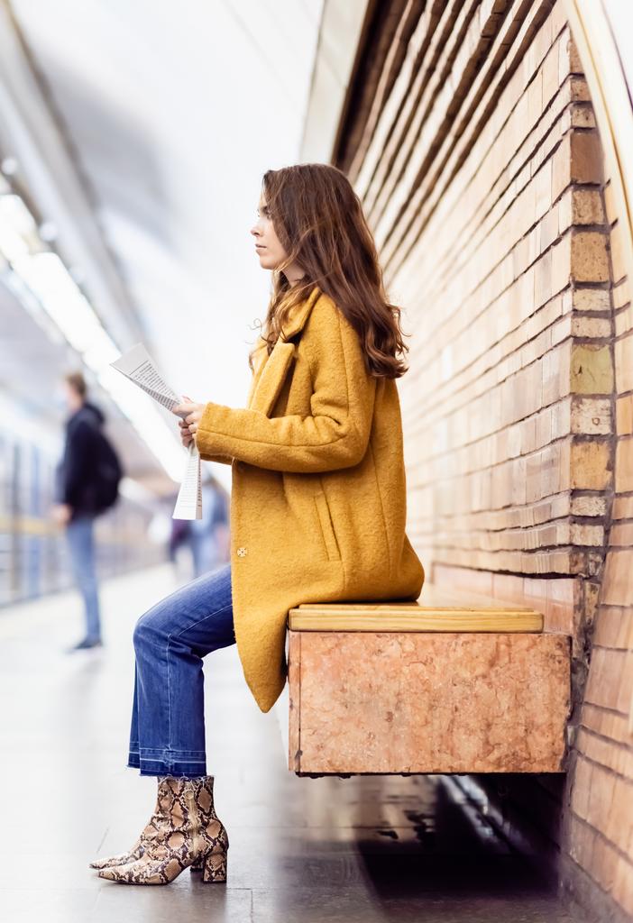 Seitenansicht einer Frau in stylischer Herbstkleidung, die Zeitung hält, während sie auf der Bank am U-Bahnsteig sitzt - Foto, Bild