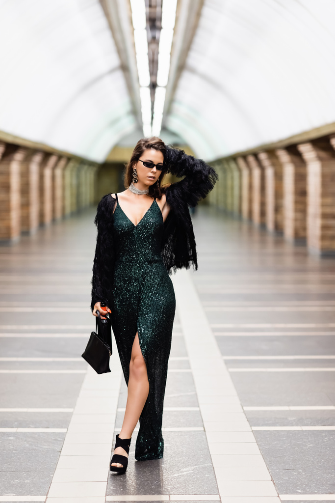 stylish woman in long lurex dress and sunglasses touching hair while posing at subway station - Fotografie, Obrázek