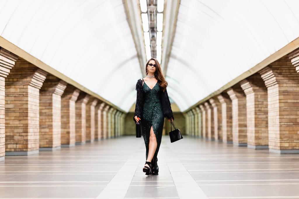 femme séduisante en robe noire longue marchant le long de la station de métro avec bouteille de vin et sac à main - Photo, image
