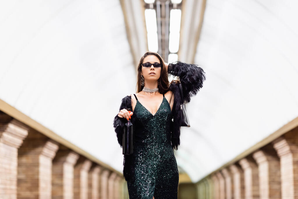 trendy woman in black lurex dress and sunglasses posing at underground station with bottle of wine - Foto, Imagen