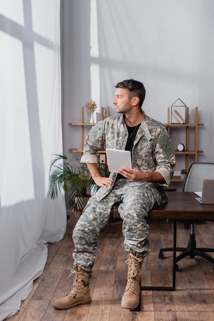 soldado en uniforme militar sosteniendo tableta digital cerca del ordenador portátil en el escritorio - Foto, Imagen