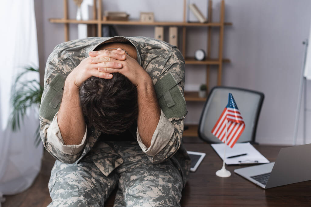militar sentado en el escritorio e inclinado cerca de gadgets y bandera americana - Foto, imagen