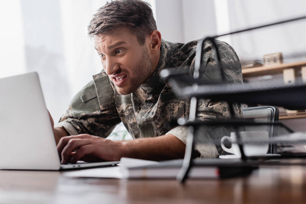 раздраженный военный, печатающий на клавиатуре ноутбука рядом с подносом для документов на размытом переднем плане - Фото, изображение