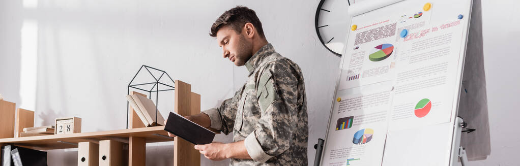 poważny wojskowy trzyma notatnik w pobliżu drewnianego stojaka i flipchart, sztandar - Zdjęcie, obraz
