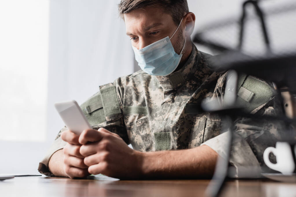 smartfon w rękach wojskowego w mundurze i masce medycznej w pobliżu tacki z dokumentami na niewyraźnym pierwszym planie - Zdjęcie, obraz