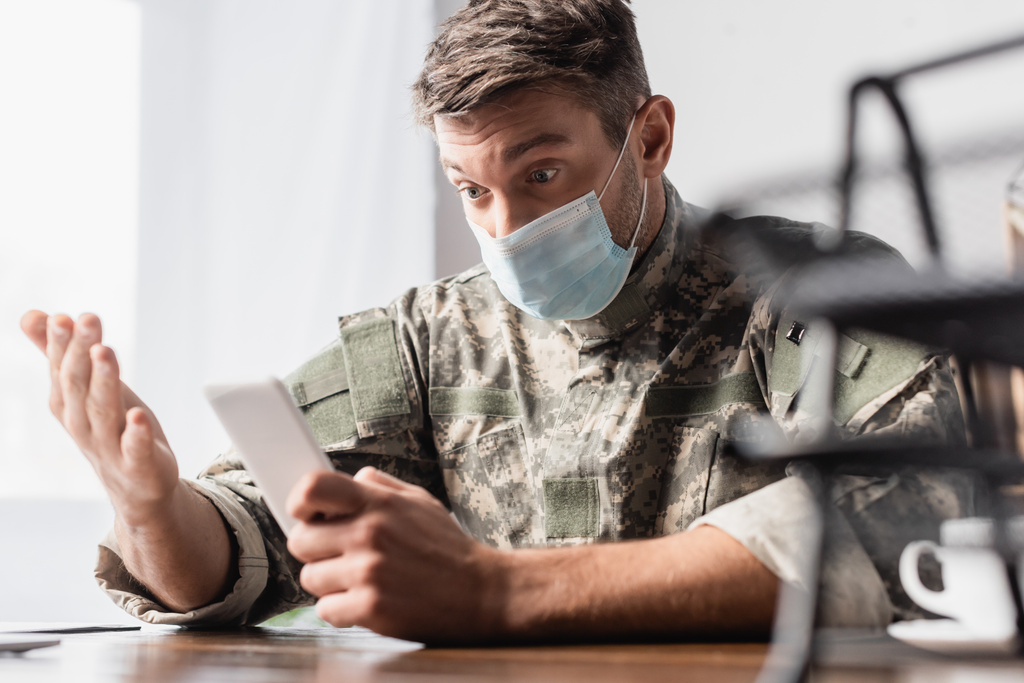 制服姿の軍人と前景がぼやけたドキュメンタリートレイの近くのスマートフォンを使った医療用マスク - 写真・画像