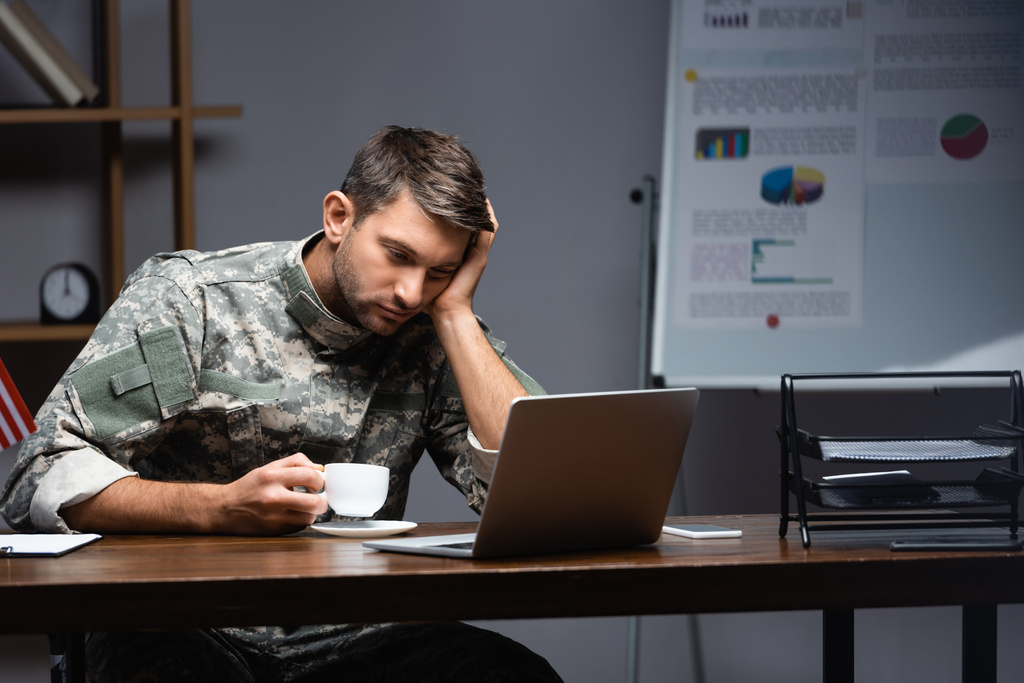 Üniformalı yorgun asker elinde kupa ve akşamları dizüstü bilgisayar tutuyor.  - Fotoğraf, Görsel