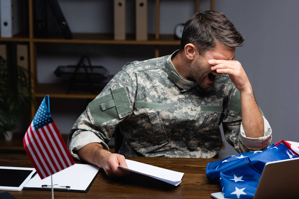 molesto militar en uniforme cubriendo los ojos y sosteniendo la carta mientras grita cerca de gadgets y bandera americana  - Foto, Imagen