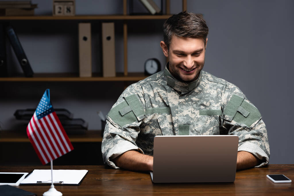homme militaire heureux en uniforme en utilisant un ordinateur portable près du drapeau américain et des gadgets  - Photo, image