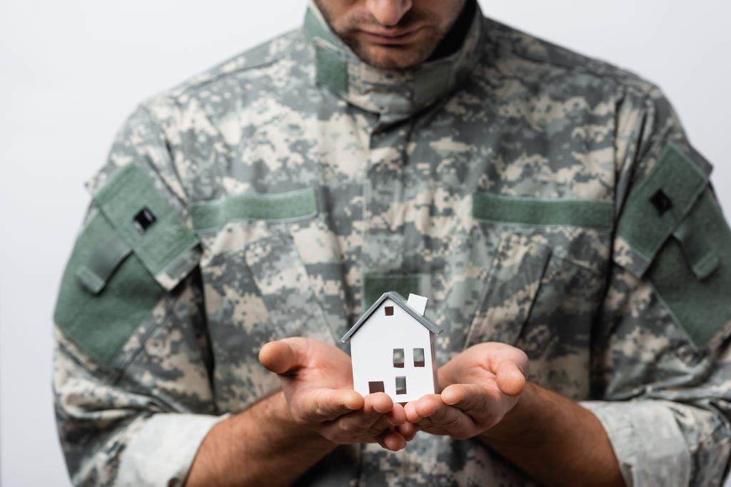 huis model in handen van patriottische militaire man in uniform op wazig achtergrond geïsoleerd op wit - Foto, afbeelding