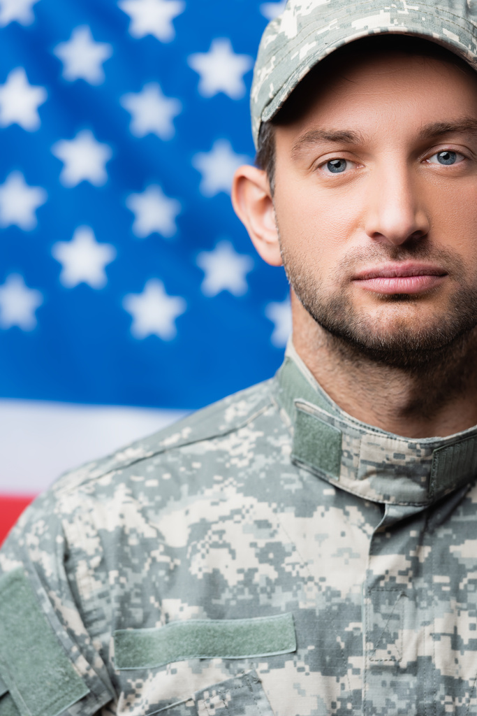 militaire man in uniform op zoek naar camera in de buurt van Amerikaanse vlag op wazige achtergrond - Foto, afbeelding