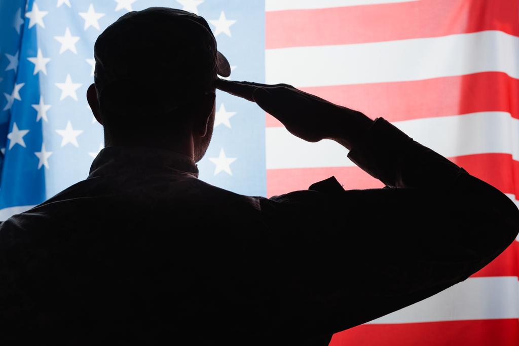 achteraanzicht van patriottische militaire man in uniform en pet geven saluut in de buurt van Amerikaanse vlag  - Foto, afbeelding