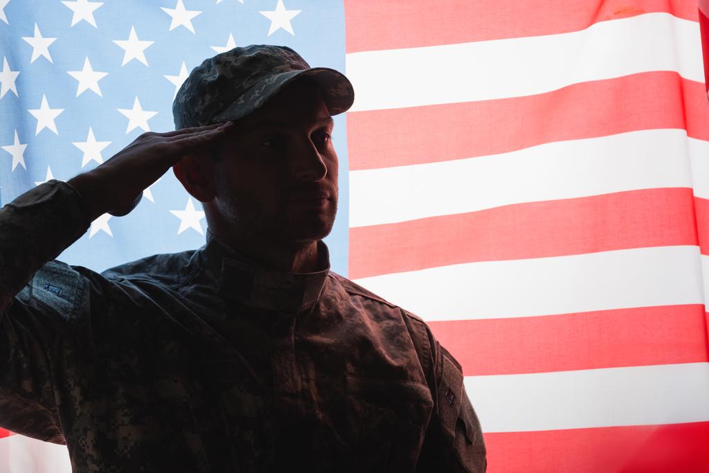 patriotic military man in uniform and cap giving salute near american flag on background - Photo, Image