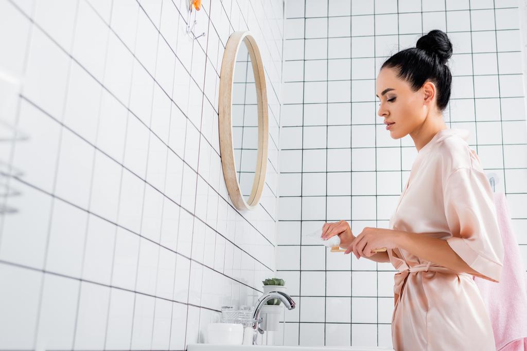 Widok z boku brunetka kobieta trzyma pastę do zębów i szczoteczkę do zębów w pobliżu lustra w łazience  - Zdjęcie, obraz