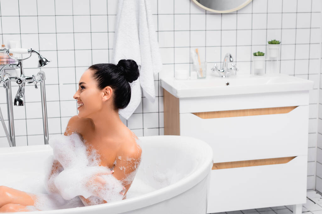 Brunette vrouw glimlachen terwijl zitten in bad met schuim  - Foto, afbeelding