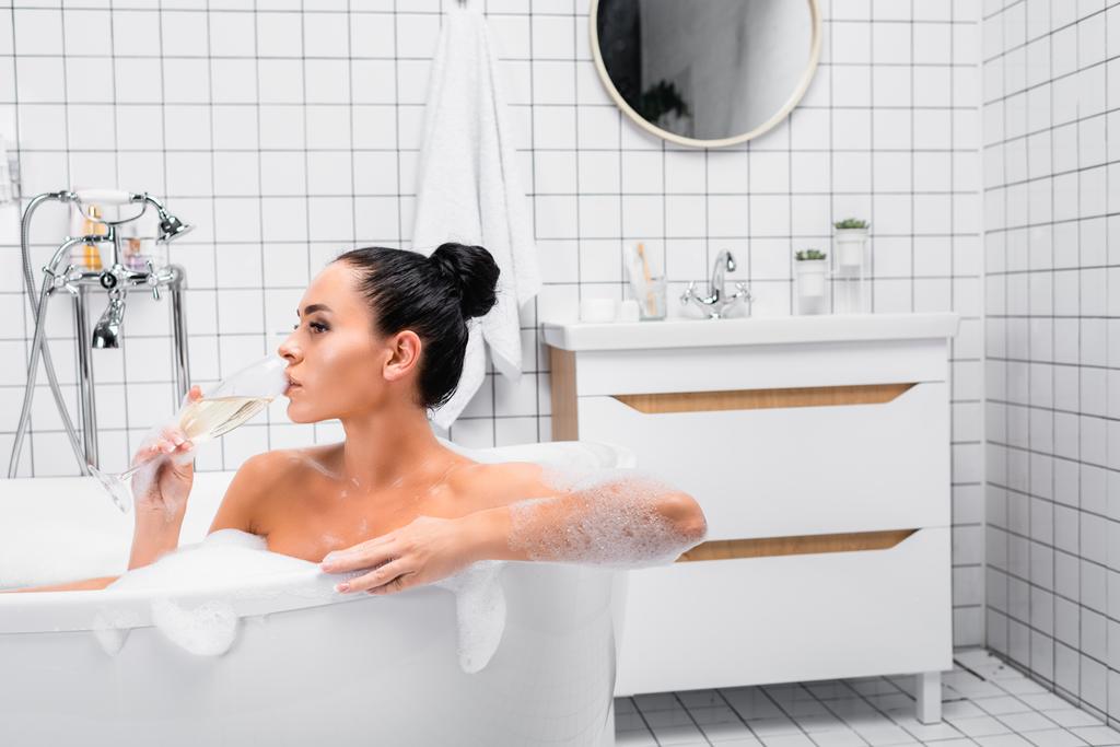 Mujer joven bebiendo champán mientras está sentada en la bañera con espuma  - Foto, imagen