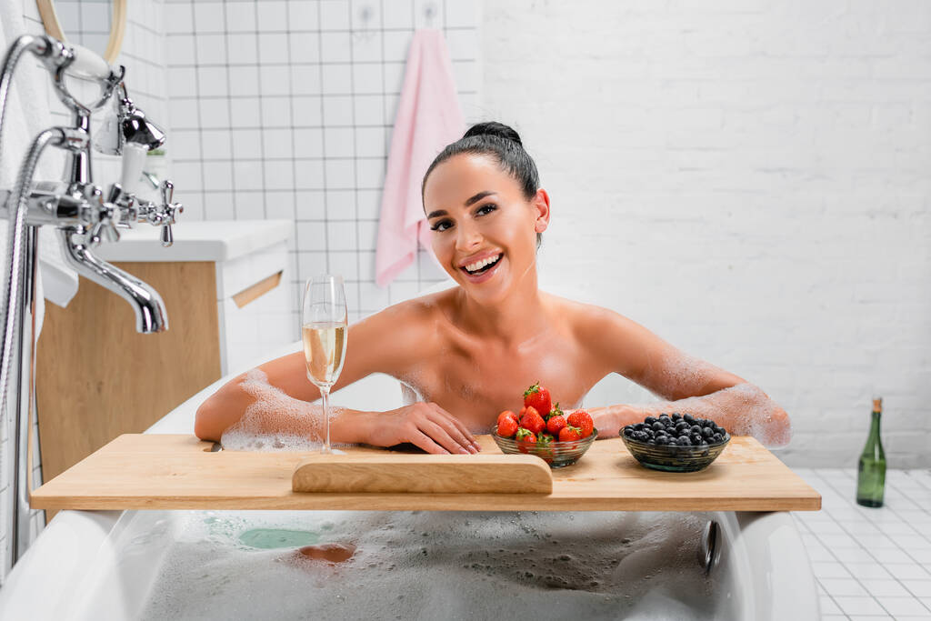 Donna allegra seduta nella vasca da bagno con schiuma vicino champagne e bacche sul vassoio  - Foto, immagini