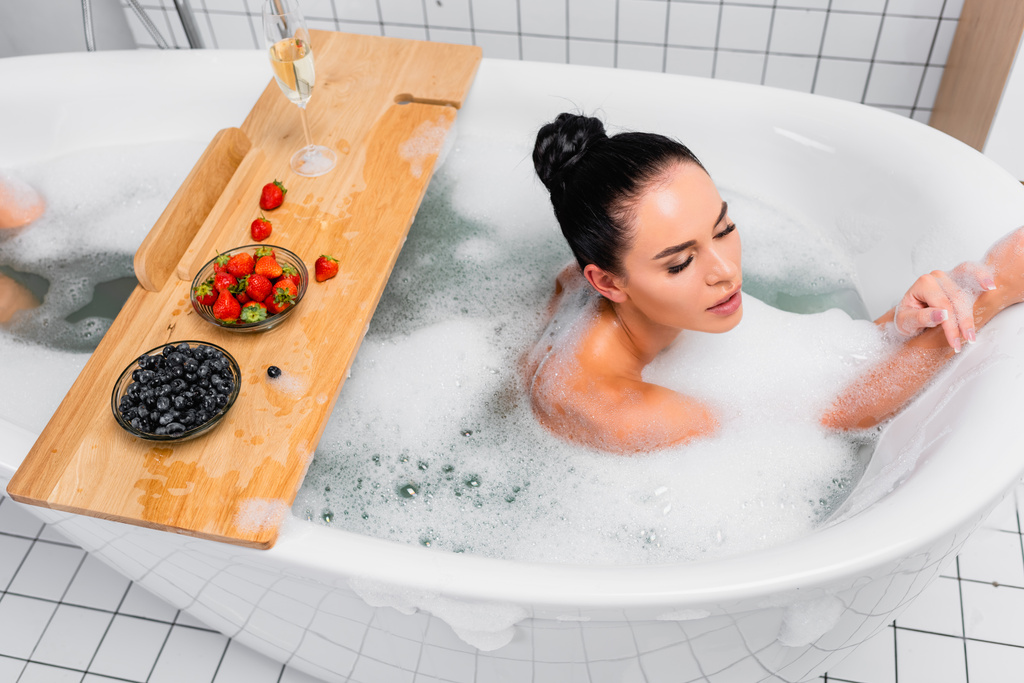 Brunette woman taking bath with foam near berries and champagne on wooden tray  - Photo, Image