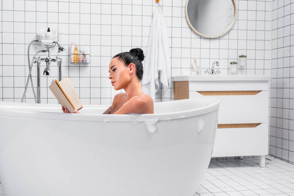 Mulher morena ler livro enquanto relaxa no banho com sabonetes em casa  - Foto, Imagem