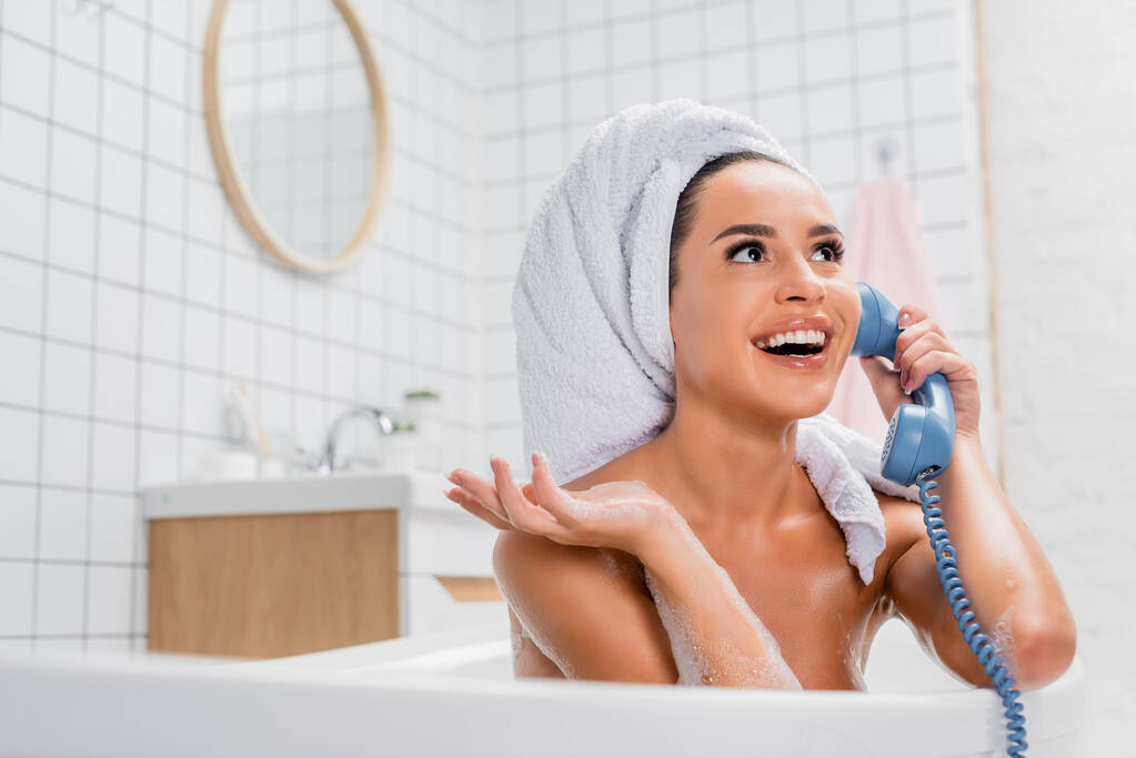 Wesoła kobieta w piance i ręcznik na głowie rozmawiająca przez telefon w wannie na niewyraźnym pierwszym planie  - Zdjęcie, obraz