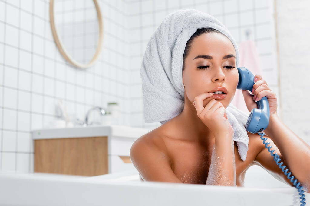 Junge Frau mit Handtuch auf dem Kopf und Finger in der Nähe der Lippen beim Telefonieren in der Badewanne im unscharfen Vordergrund  - Foto, Bild