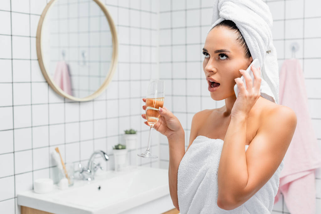 Geschockte Frau in Handtüchern spricht auf Smartphone und hält Glas Champagner im Badezimmer vor verschwommenem Hintergrund  - Foto, Bild