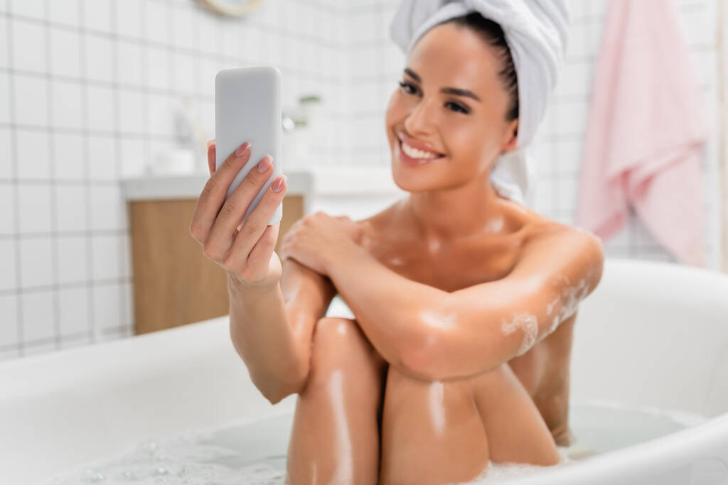 Smartphone en mano de mujer sonriente en toalla tomando selfie en baño sobre fondo borroso  - Foto, imagen