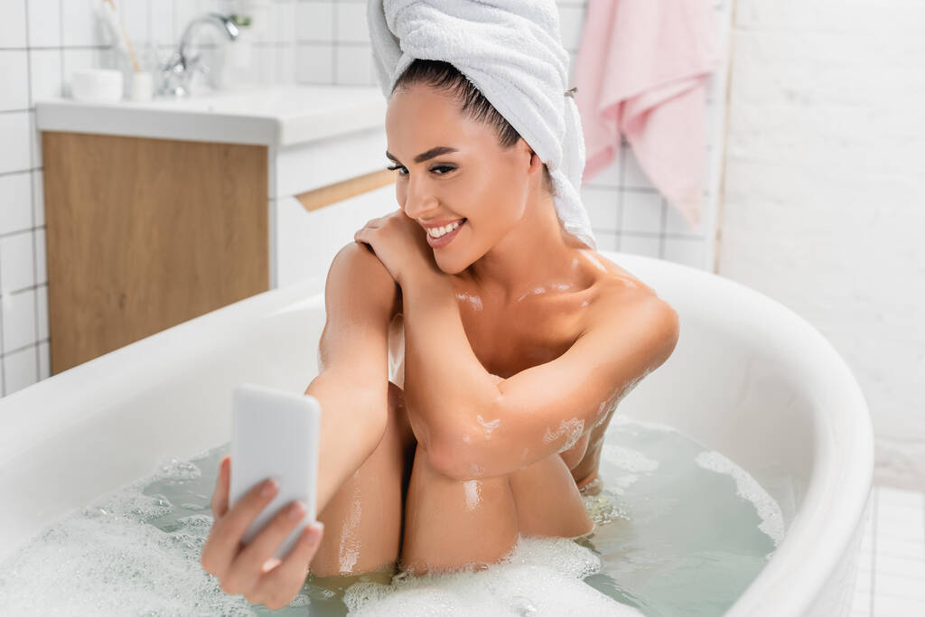 Mujer alegre tomando selfie en el teléfono inteligente en primer plano borrosa en el baño  - Foto, imagen