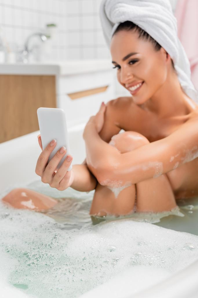 Smartphone en la mano de la mujer joven en la toalla tomando selfie en el teléfono inteligente sobre fondo borroso en el baño  - Foto, imagen