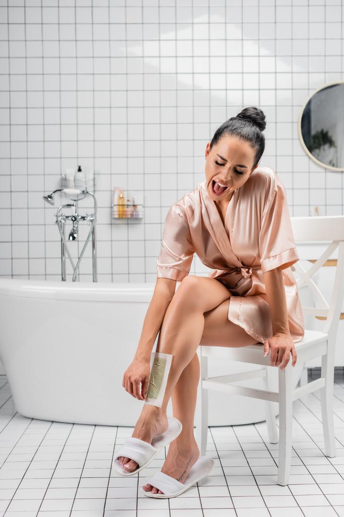 Femme hurlante en peignoir de soie décoller bande d'épilation dans la salle de bain  - Photo, image