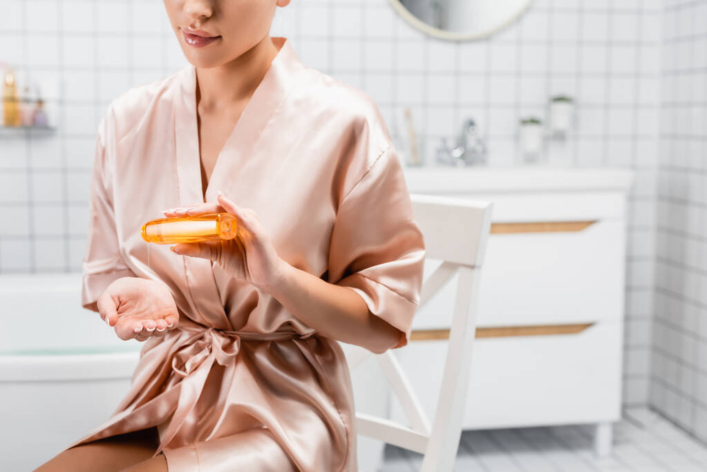 Ausgeschnittene Ansicht einer Frau im seidenen Bademantel, die Kosmetiköl ins Badezimmer gießt  - Foto, Bild