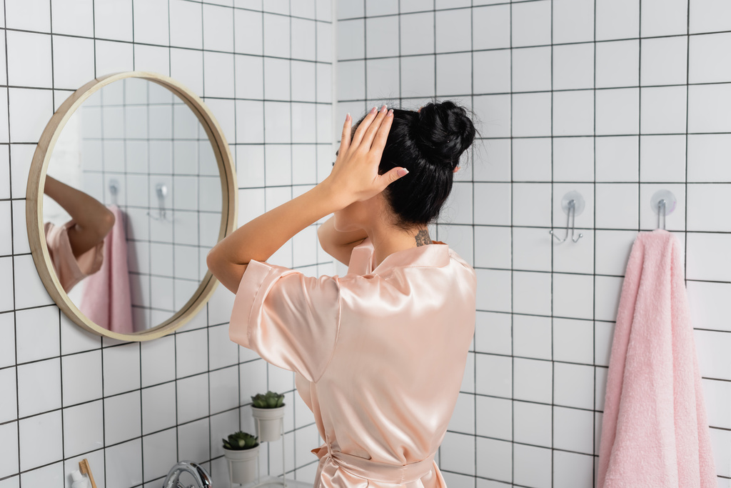 Νεαρή γυναίκα σε σατέν μπουρνούζι αγγίζοντας τα μαλλιά κοντά στον καθρέφτη στο μπάνιο  - Φωτογραφία, εικόνα
