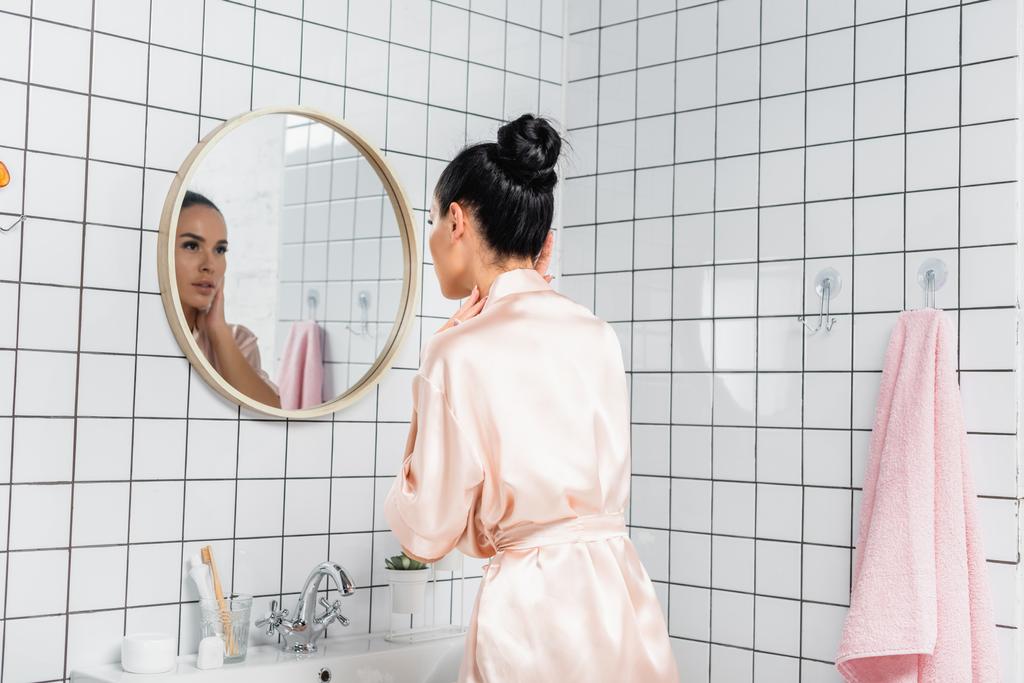 Молодая женщина в атласном халате касается кожи возле зеркала в ванной комнате - Фото, изображение