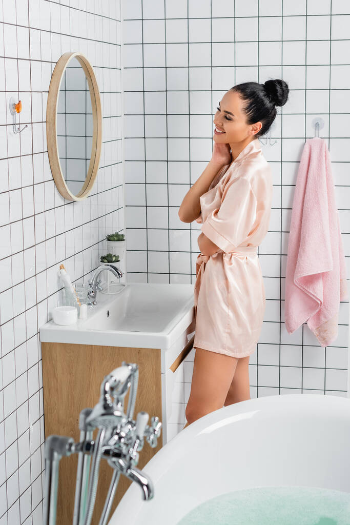 Mujer alegre en albornoz de seda mirando el espejo cerca de la bañera en primer plano borroso  - Foto, Imagen