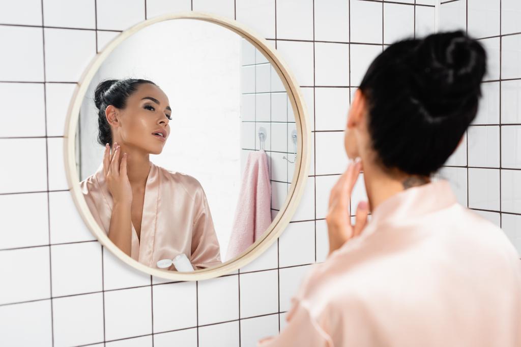 Nuori nainen soveltamalla kosmeettinen kerma kaulan lähellä peili kylpyhuoneessa  - Valokuva, kuva