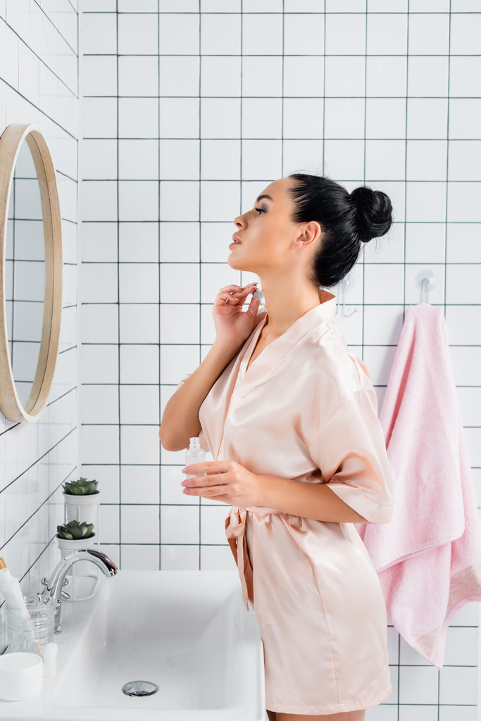 Young woman in silk bathrobe applying cosmetic serum near mirror in bathroom  - Photo, Image